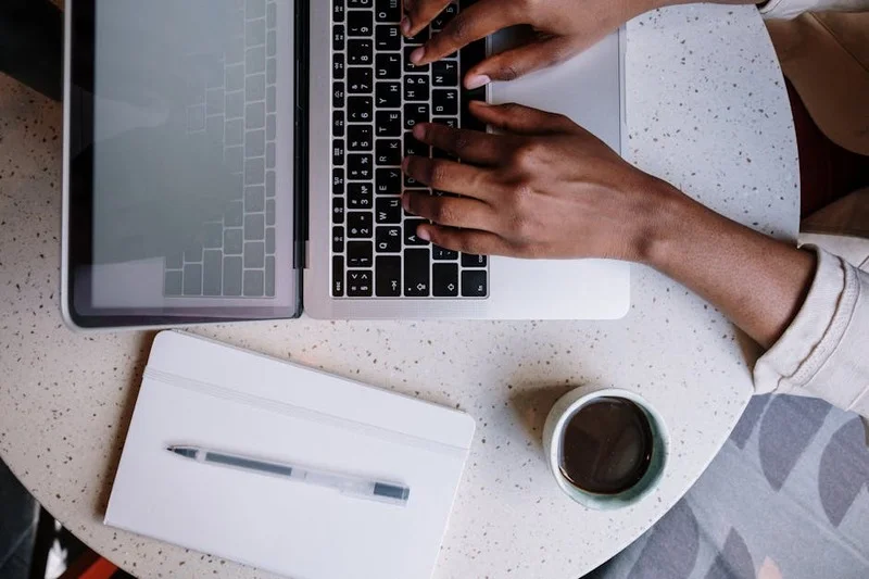 Manos de persona tecleando en teclado con cuaderno y bolígrafo para planificar en cafetería luminosa